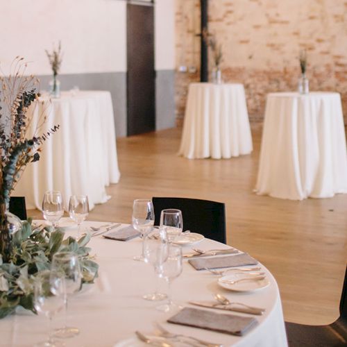 A banquet setup with round tables, white tablecloths, glassware, and greenery centerpieces in a bright event hall.