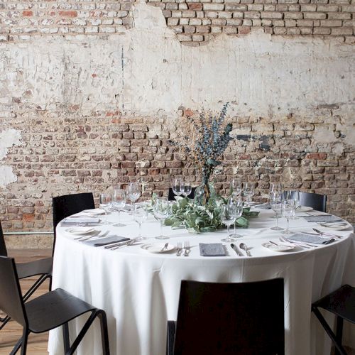 A round dining table with a white tablecloth, set with silverware and glassware, surrounded by black chairs in a rustic brick-walled room.
