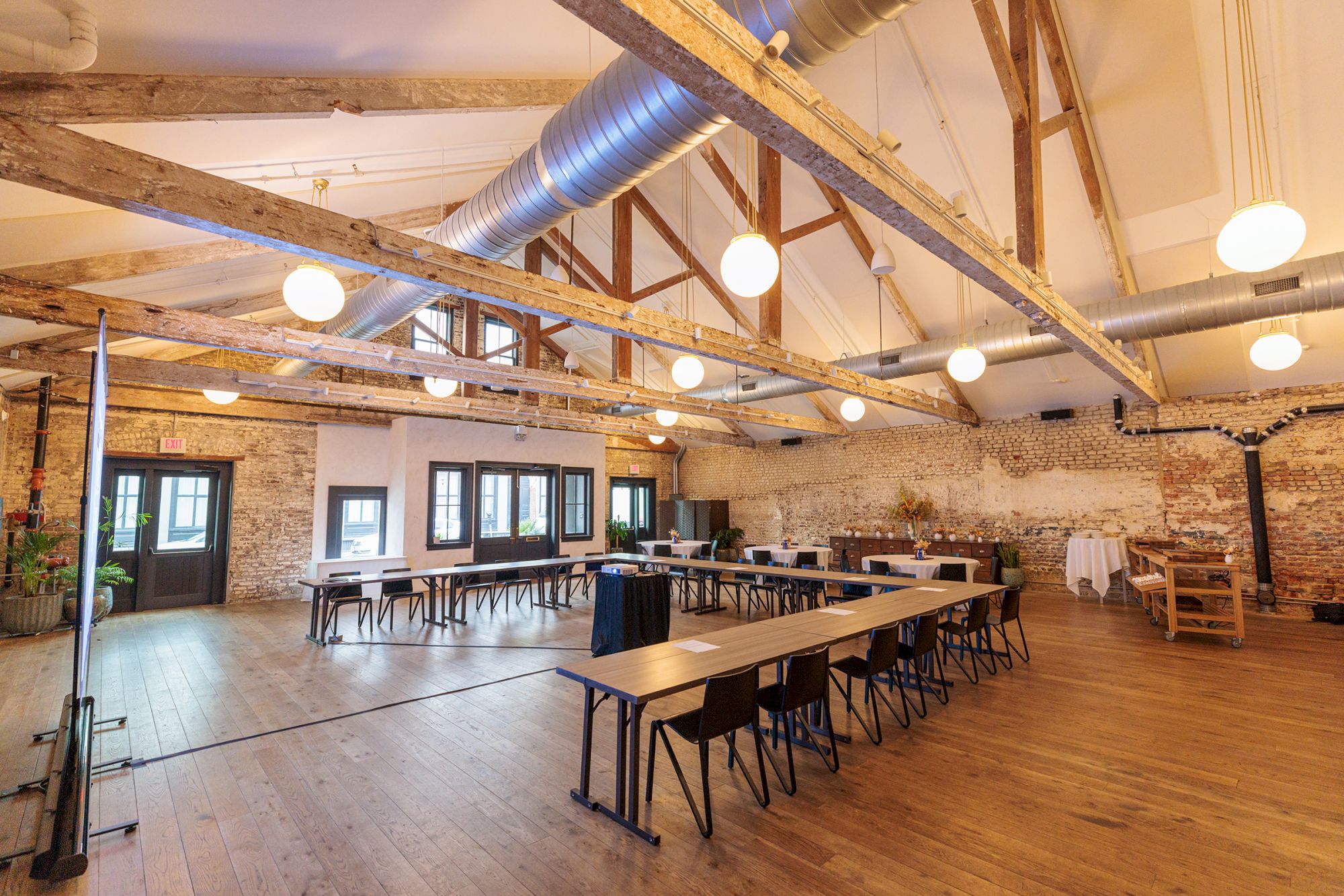 A bright, industrial-style event space with exposed brick walls, long tables, scattered chairs, wooden floors, and overhead lighting.